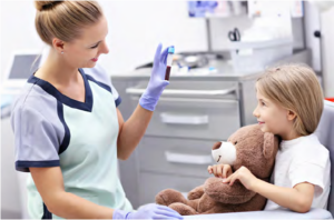 Photo of a healthcare worker sitting next to a female child holding a teddy bear.